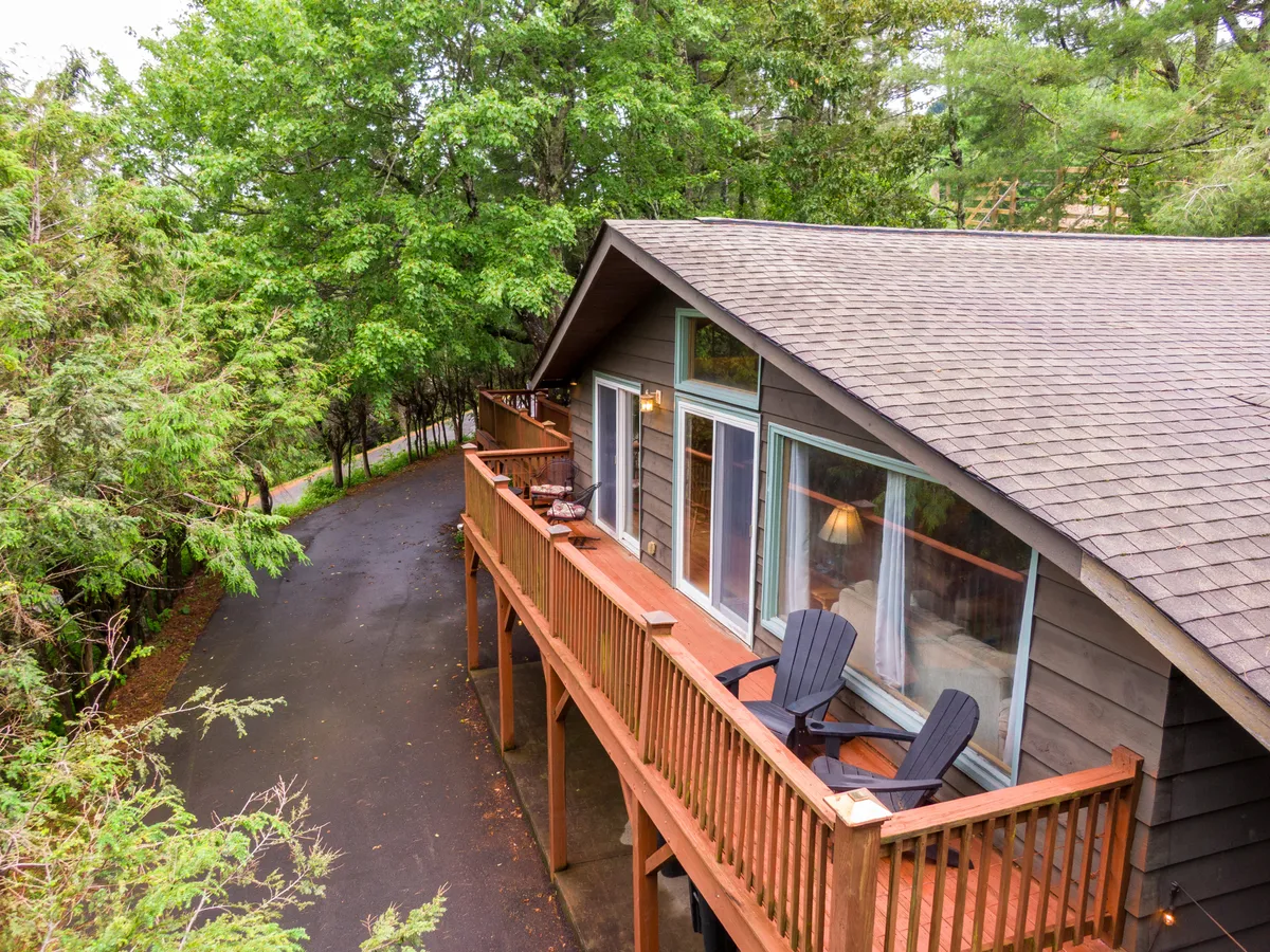 A tranquil wooden deck extends from the cabin, furnished with Adirondack chairs and surrounded by lush greenery. Large windows reflect the sun, and the deck invites guests to relax and enjoy the mountain air. The setting hints at both sunrise coffee and sunset conversations, seamlessly blending indoor comfort with outdoor beauty.