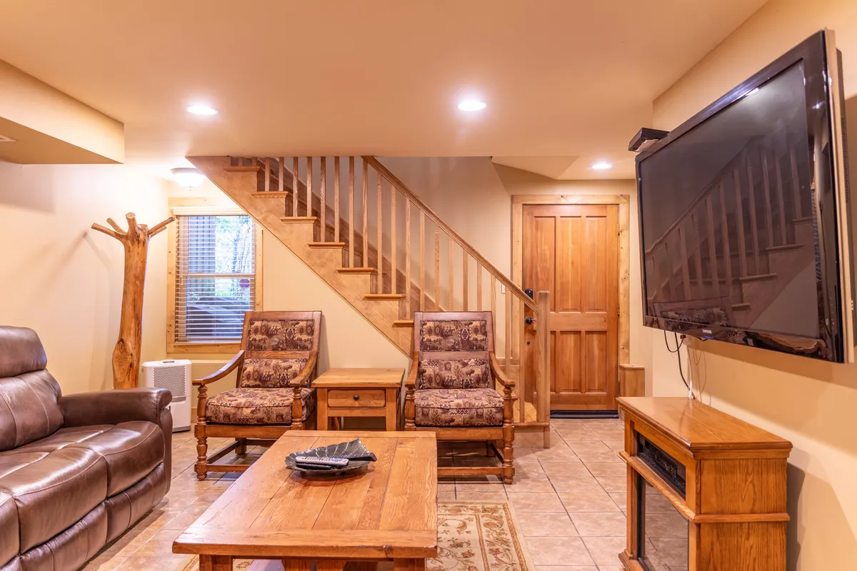 A spacious and welcoming living area furnished with a leather sofa, wooden coffee table, TV, armchairs, and a staircase leading to other parts of the home. The layout encourages socializing, relaxing, and group activities.