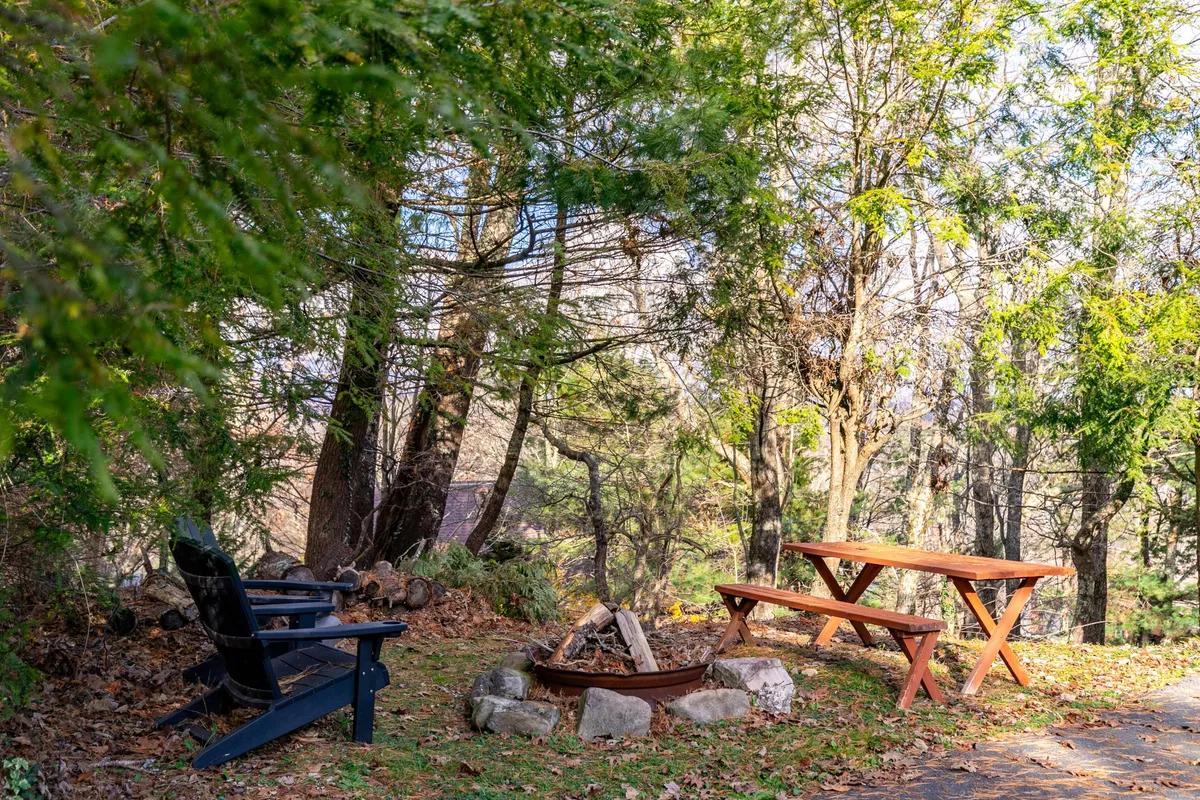 An exterior scene featuring a stone fire pit surrounded by adirondack chairs and a picnic table. The setting is nestled in nature, ideal for gathering with friends and family under the open sky. The fire pit serves as a central focal point, promising cozy evenings outdoors in every season.