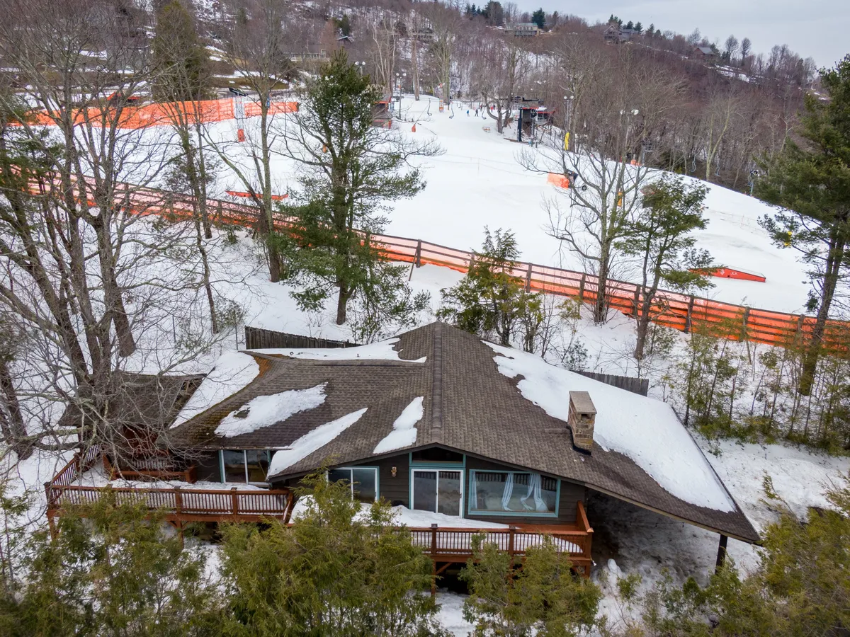 The image shows a striking A-frame cabin nestled in a snowy landscape on Appalachian Ski Mountain, with the cozy home standing out against the tranquil wintry backdrop. The surrounding snow and nearby orange fencing hint at direct access to winter sports and play, while the soft light suggests warmth and comfort inside.