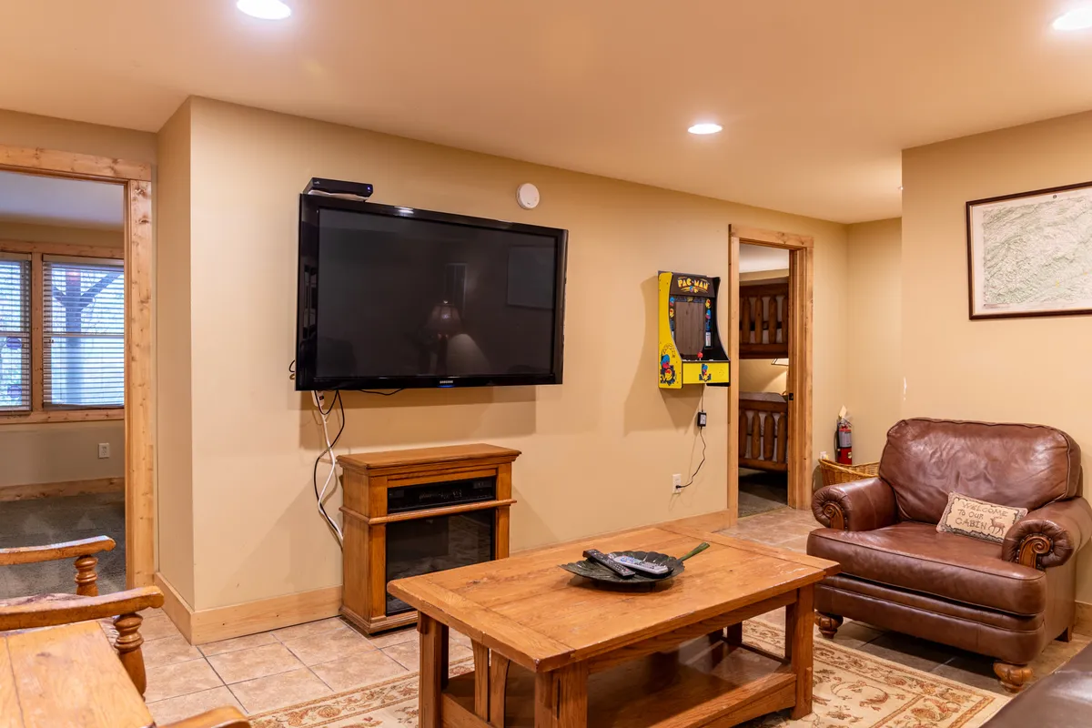 A cozy entertainment room featuring a prominent Pacman arcade game machine, a large television, a wooden coffee table, and a comfortable leather armchair set against a warm, inviting backdrop. Soft lighting creates an atmosphere ideal for gaming, movie nights, and family gatherings.