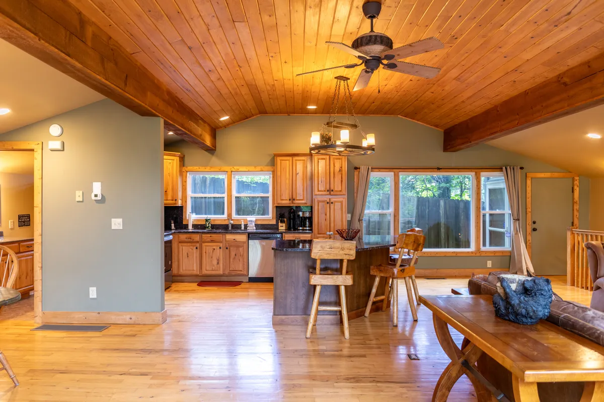 A wide view of the dining area and kitchen, showcasing a wooden ceiling, kitchen cabinetry, a large window filling the space with natural light, and a connected dining table—emphasizing the open-concept design and scenic surroundings.