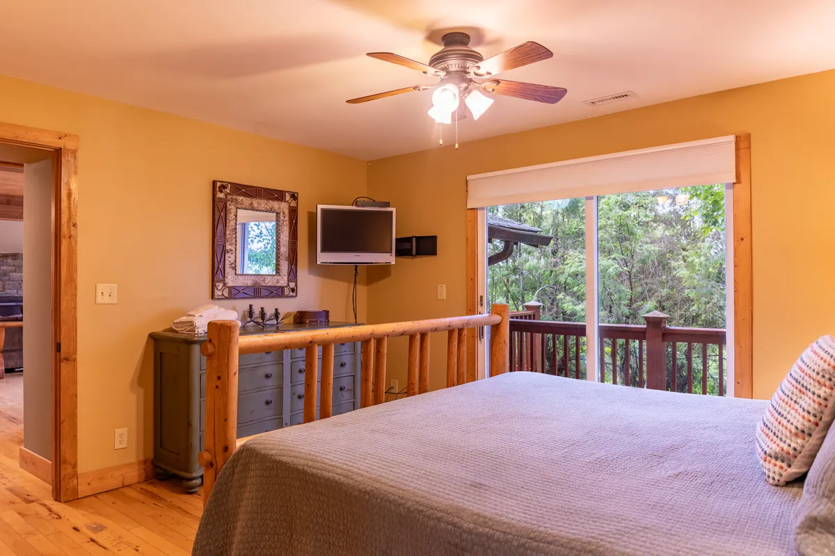A cozy bedroom with a rustic log frame bed, a large window offering a view of the outdoors, a decorative mirror, and a television. Soft lighting enhances the inviting, tranquil atmosphere.
