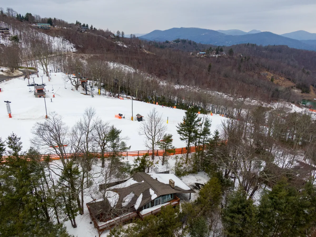 This image captures the house in the foreground with a backdrop of snow-covered ski slopes and surrounding trees. The tranquil scene includes people skiing and snowboarding, evoking the quintessential slopeside winter experience just steps from the property.