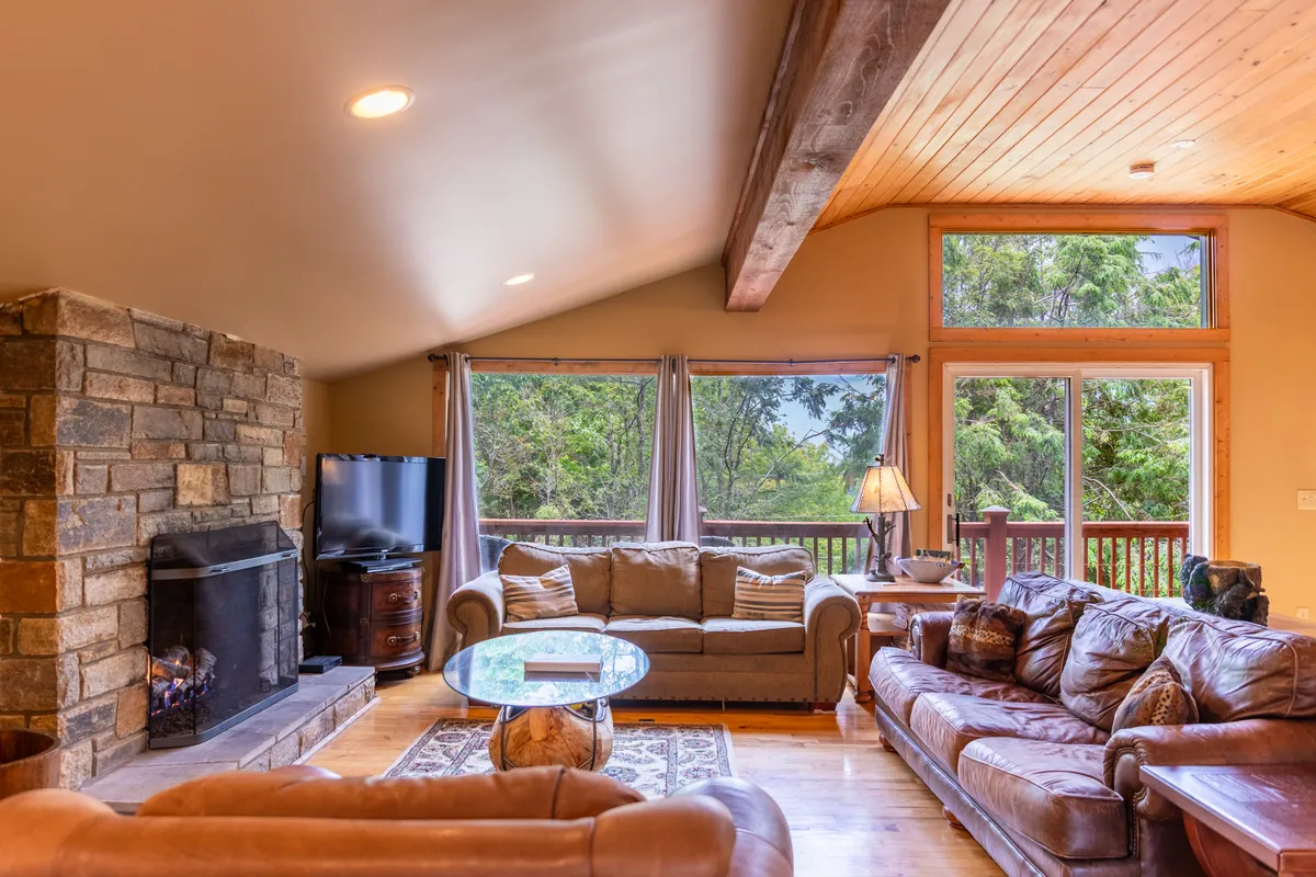 This image features a welcoming great room with a stone fireplace, large windows framing expansive mountain views, plush sofas, and a glass coffee table. The natural light streaming in enhances the warmth of the comfortable, open space, and the arrangement is ideal for gatherings and relaxation.